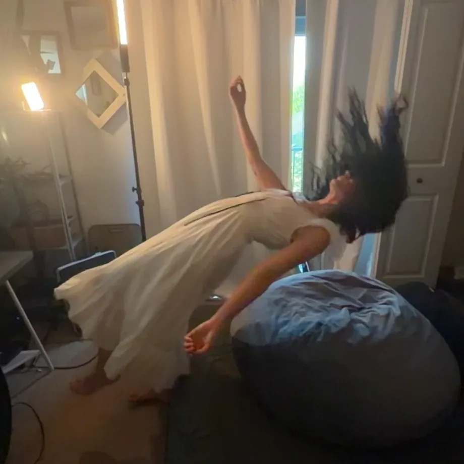 Jessie bending backwards in a white dress at home, hair flying, lamplight and curtains behind her